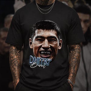 May include: A black short-sleeved t-shirt featuring a large graphic print of the face of boxer Dmitry Bivol. The design includes the text "BIVOL" on the teeth, "DMITRY" in a blue stylized font, and the name "Bivol" in a white cursive script below.