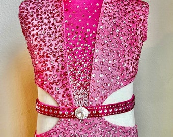 May include: A pink dance leotard with a high neckline and a mesh panel in the front. The leotard is covered in rhinestones and has a cutout at the waist. It has a pink belt with a large rhinestone buckle. The leotard is on a white mannequin and has a tag that says "SZ 8/10".
