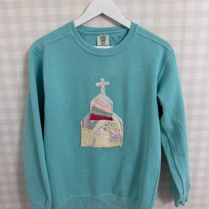May include: A turquoise crew neck sweatshirt with a church-shaped patch on the front. The patch is made from fabric scraps and has a cross. The sweatshirt is hanging on a white hanger. The label reads "Comfort Colors".
