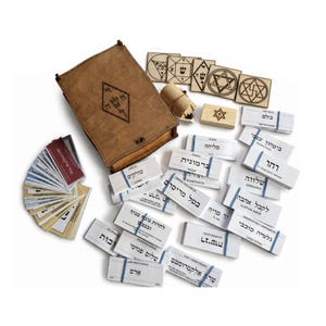 May include: A brown wooden box with a diamond-shaped symbol, a small wooden cylinder, four square wooden tiles with geometric engravings, and numerous rectangular white paper cards bundled with blue rubber bands, featuring Hebrew text.
