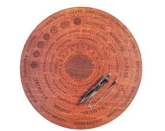 May include: A wooden divination board with engraved text and symbols, featuring a pendulum with a silver chain. The board has concentric circles with names, months, and spiritual concepts. The pendulum is black and brown.