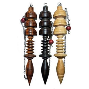 May include: Three wooden pendulum dowsing tools with different wood grain patterns. The pendulums are brown, black, and natural wood. Each pendulum has a chain with a small red bead at the end.