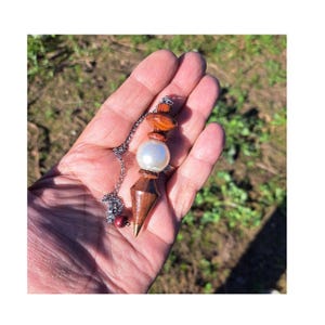 Puede incluir: Un péndulo con una cadena plateada y un diseño de madera y piedra. El péndulo presenta una base de madera en forma de lágrima, una gran perla blanca y varias piedras naranjas y marrones. El péndulo se sostiene en una mano.