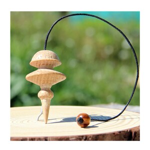 May include: A light-colored wooden pendulum with a tiered, rounded top and a pointed base. A thin black cord is attached to the top, curving downward to connect to a small, polished brown wooden bead resting on the surface below the pendulum.