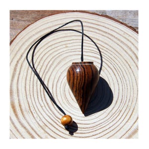 May include: A teardrop-shaped wooden pendulum featuring a dark brown and black striped grain pattern. The pendulum is attached to a thin black cord that includes a small, smooth, light brown wooden bead near the end of the string.
