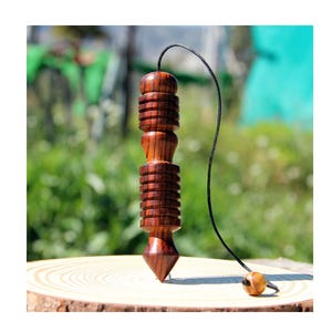 Può includere: Un pendolo in legno marrone scuro con un corpo cilindrico caratterizzato da scanalature orizzontali e una punta affilata. Il pendolo è sospeso da un sottile cordino nero che termina con una piccola perla di legno rotonda e chiara.