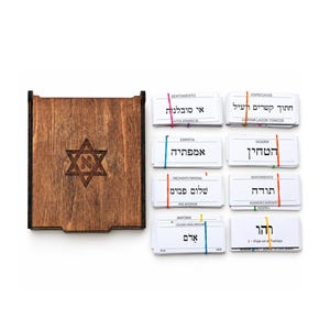 May include: A dark brown wooden box with a Star of David engraving containing the Hebrew letter Aleph, accompanied by eight stacks of white rectangular cards bound with colorful rubber bands, each featuring Hebrew text and Spanish headings.