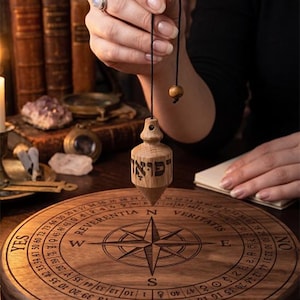 May include: A wooden pendulum with Hebrew text suspended over a round wooden board featuring a compass rose, numbers, and the words YES, NO, REVERENTIA, and VERITAS. The board includes additional Hebrew text and astrological symbols.