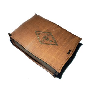 Op de afbeelding: Een lichtbruine houten kist met een ruitvormig ontwerp in het midden. De ruit is een lichtere bruintint en heeft een geometrisch patroon. De kist heeft een zwarte rand en een kleine 'B' aan de zijkant.