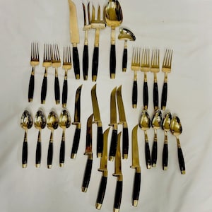 May include: A collection of vintage flatware with gold-toned metal and black handles. The set includes knives, forks, spoons, and serving utensils. The arrangement is on a white surface, showcasing the complete set.