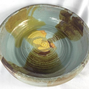 May include: A large, round ceramic bowl with a blue-green glaze. The bowl has a yellow and brown center with a concentric circle pattern. The bowl has a rustic, handmade look.