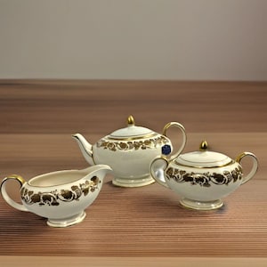May include: A three-piece tea set with a white base and gold floral detailing. The set includes a teapot, a sugar bowl with a lid, and a creamer. The teapot and sugar bowl have gold handles and finials. The set is displayed on a wooden surface.