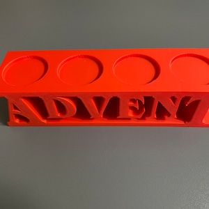 Op de afbeelding: Fel oranje 3D-geprinte adventskalender met vier cirkelvormige uitsparingen en het woord "ADVENT" uitgesneden. Een decoratief en praktisch item om af te tellen.