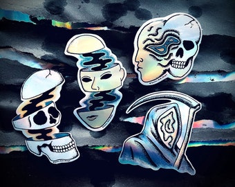 May include: Four iridescent stickers featuring black line drawings of skulls and faces. The stickers are displayed against a dark grey background with a holographic sheen.