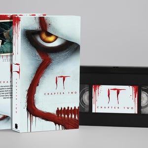 Könnte beinhalten: Eine weiße VHS-Hülle mit dem Titel IT CHAPTER TWO in rotem Text, einem großen gelben Auge und einer roten Liniengrafik mit Silhouetten. Ein schwarzes VHS-Band ist enthalten, mit einem weißen Etikett, das das rote IT CHAPTER TWO Logo zeigt.
