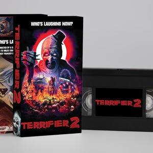 May include: A black VHS tape and two VHS cases for the horror film "Terrifier 2." The cases feature the film's title and artwork, with the text "Who's Laughing Now?" The tape itself also displays the film's title in red.