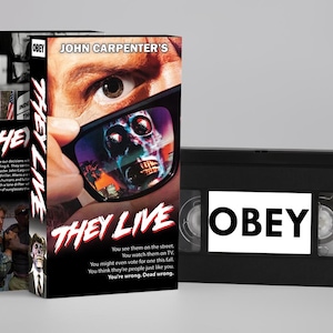 Könnte beinhalten: Eine VHS-Kassette und Box-Set für den Film "They Live". Das Box-Art zeigt ein Auge und eine Sonnenbrille, die einen Schädel reflektiert. Auf der VHS-Kassette steht "OBEY".