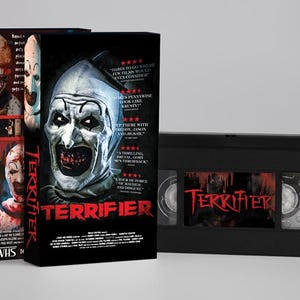 Könnte beinhalten: Eine schwarze VHS-Kassette und zwei VHS-Kassettenhüllen für den Horrorfilm "Terrifier". Die Hüllen zeigen einen bedrohlichen Clown mit weißer Schminke und einem blutigen Mund. Die VHS-Kassette selbst trägt den Filmtitel in Rot.