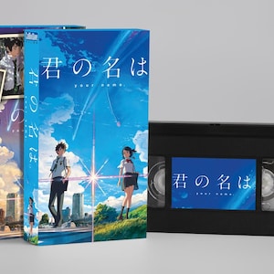 Op de afbeelding: Een blauwe VHS-band en twee bijpassende dvd-hoesjes met anime-kunstwerk. De hoesjes en de band hebben de titel "Your Name" in het Japans. De hoesjes tonen personages in een schilderachtige omgeving met een sterrenhemel.