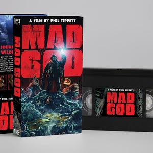 Op de afbeelding: VHS-tape en boxset voor de film "Mad God". De box art toont een figuur op een rotsachtig landschap met de titel in grote rode letters. De VHS-tape is zwart met de filmtitel op het label.