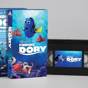 Op de afbeelding: Twee VHS-banden en een VHS-bandhoes voor de Disney Pixar-film "Finding Dory". De banden en de hoes zijn zwart en blauw en tonen afbeeldingen van Dory, Nemo en andere personages uit de film. De titel "Finding Dory" is prominent aanwezig.