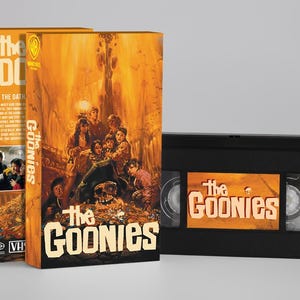 Op de afbeelding: Twee VHS-tapeboxen en een VHS-tape van de film "The Goonies". De dozen zijn oranje met de filmtitel en een groep mensen. De VHS-tape is zwart met de filmtitel op het label.