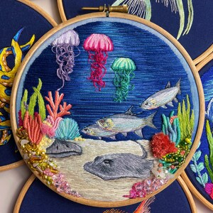 May include: Embroidered hoop art depicting an underwater scene with coral, fish, jellyfish, and a stingray. The background is a deep blue with white and light blue accents. The hoop is made of wood and has a gold screw.