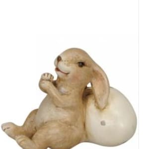 May include: A light brown bunny figurine with a cream-coloured egg. The bunny is lying down, with its paws clasped together. The egg is behind the bunny. The figurine is a decorative item.