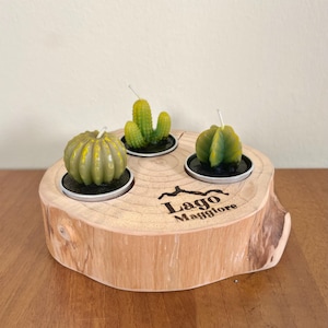 Può includere: Un portacandele in legno con tre candele a forma di cactus. Le candele sono verdi e gialle, poste in piccoli piattini neri. Il supporto è una sezione trasversale di un tronco d'albero, con il testo "Lago Maggiore".