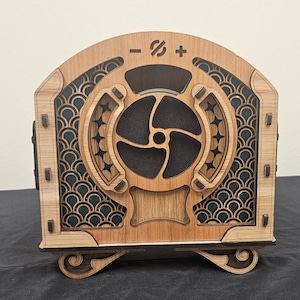 Puede incluir: Una caja de madera con un panel frontal decorativo que presenta un diseño de ventilador estilizado y los símbolos "-", "#" y "+". La caja tiene un acabado marrón oscuro y detalles intrincados.