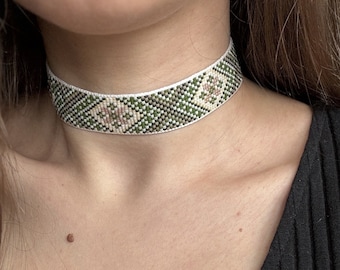 Op de afbeelding: Een kralen choker ketting met een geometrisch patroon. De choker heeft een witte basis met groene en roze ruitvormige accenten. De ketting wordt gedragen tegen een zwarte geribbelde top.