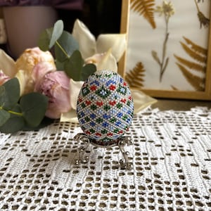 Peut inclure: Un &oelig;uf de P&acirc;ques perl&eacute; avec un motif g&eacute;om&eacute;trique dans des tons de bleu, vert, rouge et blanc. L'&oelig;uf est pr&eacute;sent&eacute; sur un support argent&eacute;.