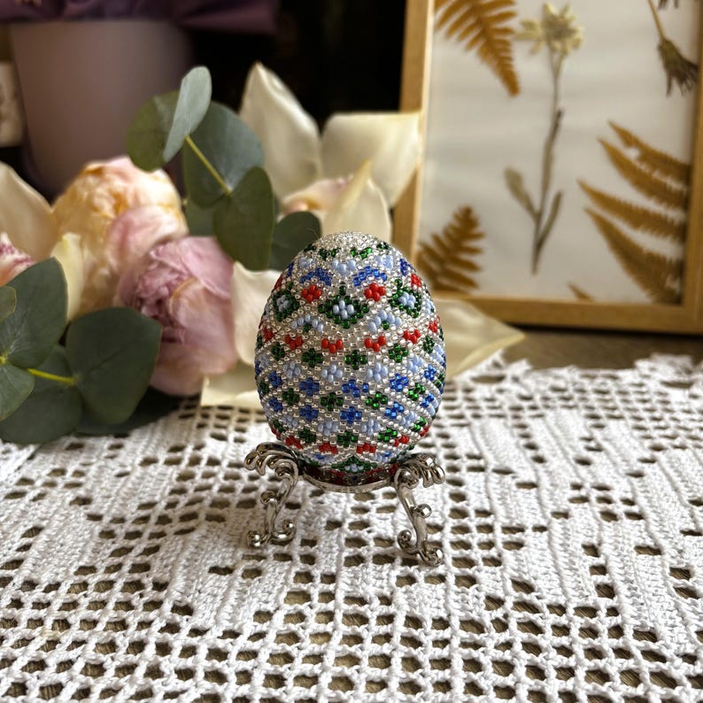 Peut inclure: Un &oelig;uf de P&acirc;ques perl&eacute; avec un motif g&eacute;om&eacute;trique dans des tons de bleu, vert, rouge et blanc. L'&oelig;uf est pr&eacute;sent&eacute; sur un support argent&eacute;.