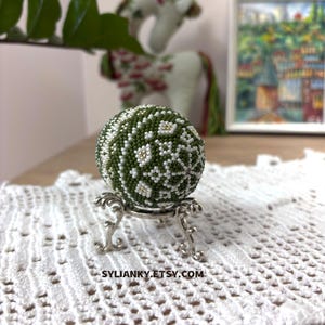Może przedstawiać: Ozdobna, koralikowa kula w odcieniach zieleni, bieli i złota, spoczywająca na srebrnym metalowym stojaku. Kula ma szczeg&oacute;łowy wz&oacute;r kwiatowy. Tło obejmuje biały koronkowy obrus i obraz.