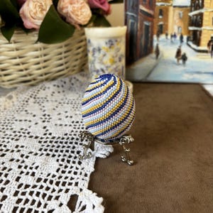 K&ouml;nnte beinhalten: Ein Perlen-Osterei mit einem gestreiften Muster in Blau, Wei&szlig;, Rosa und Gold, auf einem silbernen St&auml;nder.