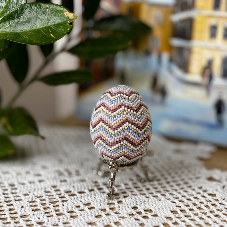 Peut inclure: Un &oelig;uf d&eacute;coratif orn&eacute; de perles en motif chevron, dans des tons de rose, bleu et or. L'&oelig;uf est pos&eacute; sur un support argent&eacute;. L'arri&egrave;re-plan est flou. Un objet d&eacute;coratif unique.