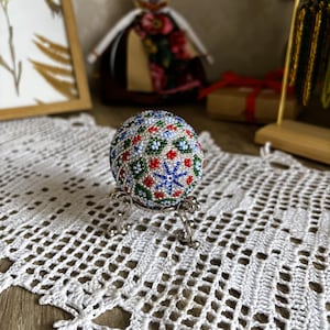 Peut inclure: Un &oelig;uf blanc avec un motif de perles color&eacute;es dans des tons de bleu, vert, rouge et blanc. L'&oelig;uf est sur un support argent&eacute;.