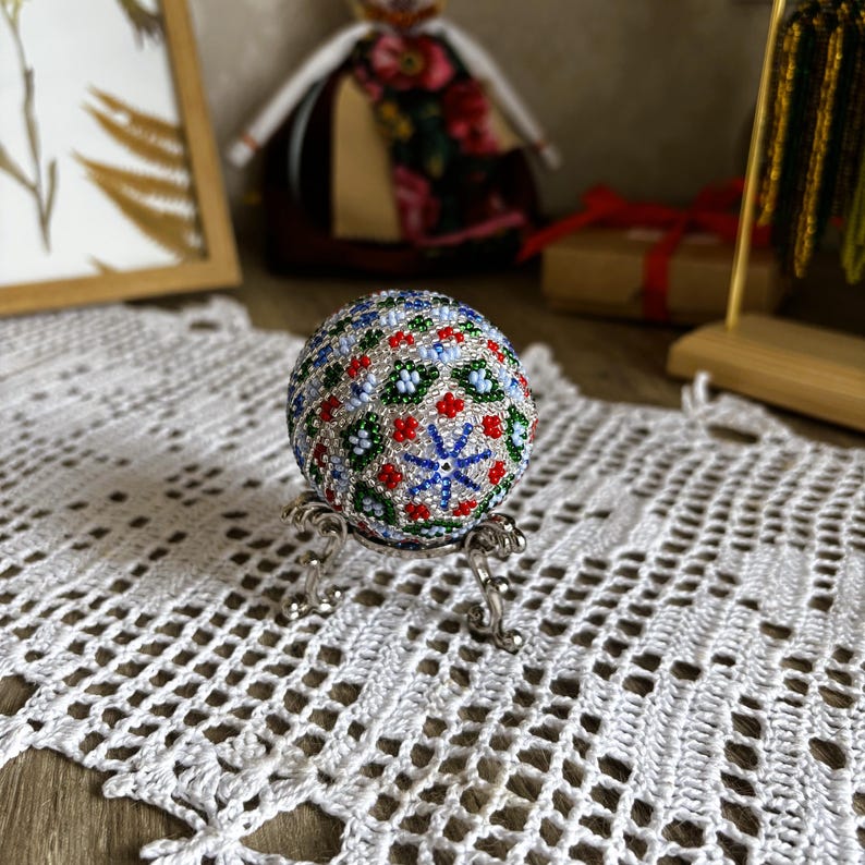 Peut inclure: Un &oelig;uf blanc avec un motif de perles color&eacute;es dans des tons de bleu, vert, rouge et blanc. L'&oelig;uf est sur un support argent&eacute;.