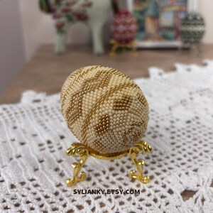 Puede incluir: Un huevo decorativo cubierto de cuentas de color crema con detalles dorados, exhibido sobre un soporte dorado ornamentado. El huevo presenta un patr&oacute;n geom&eacute;trico. El nombre de la tienda Etsy, SYLIANKY.ETSY.COM, es visible en la parte inferior.