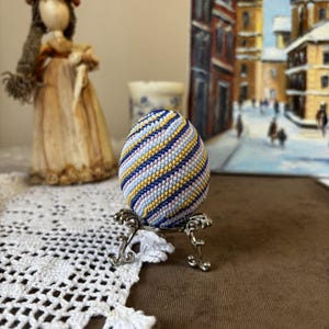 K&ouml;nnte beinhalten: Ein Perlen-Osterei mit einem Spiralmuster in Blaut&ouml;nen, Gelb, Rosa und Wei&szlig;. Das Ei wird auf einem silbernen St&auml;nder pr&auml;sentiert.