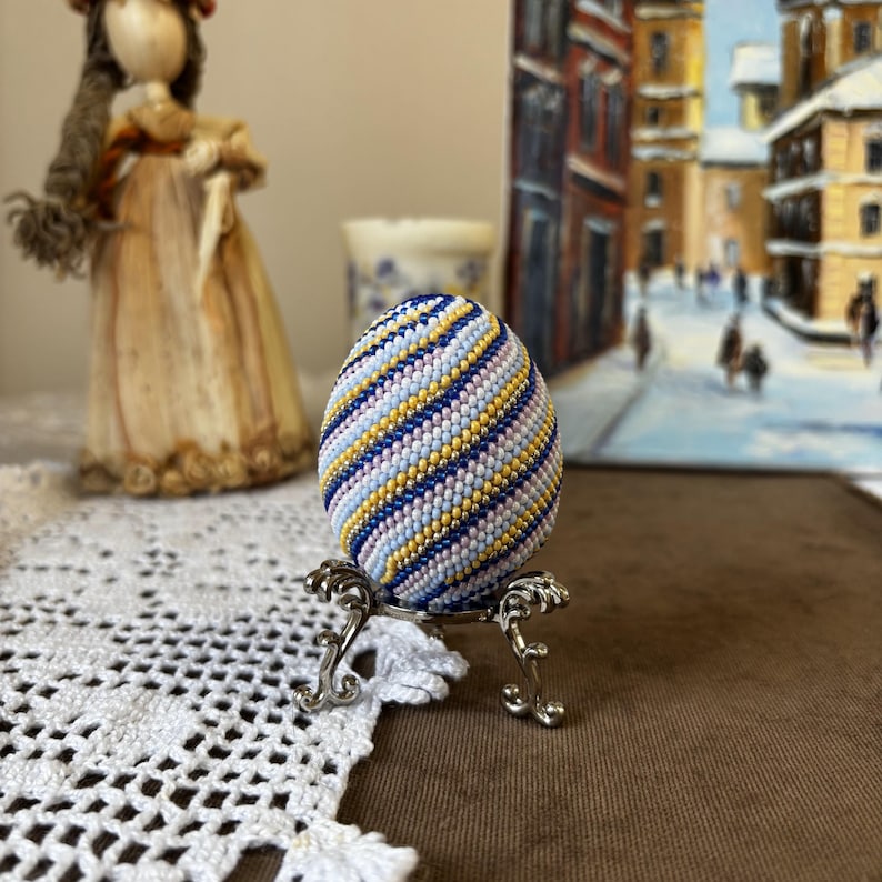 K&ouml;nnte beinhalten: Ein Perlen-Osterei mit einem Spiralmuster in Blaut&ouml;nen, Gelb, Rosa und Wei&szlig;. Das Ei wird auf einem silbernen St&auml;nder pr&auml;sentiert.