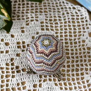 Peut inclure: Une sph&egrave;re perl&eacute;e d&eacute;corative aux tons pastel de bleu, rose et or, pos&eacute;e sur un support argent&eacute;. La surface de la sph&egrave;re est orn&eacute;e d'un motif de perles d&eacute;taill&eacute;. Le fond est un tissu blanc &agrave; tissage ouvert.