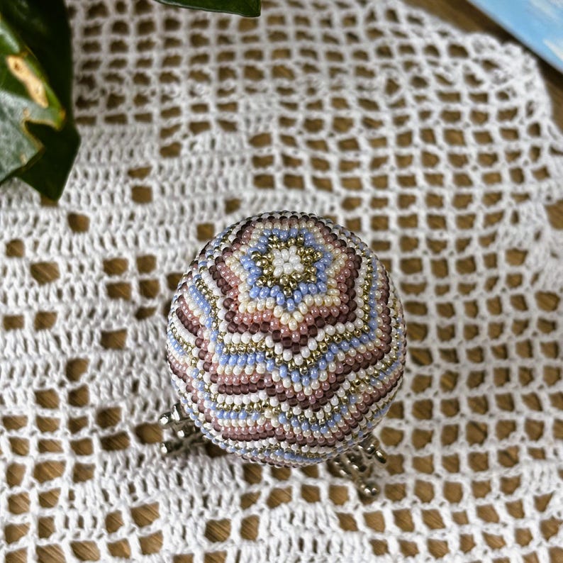 Peut inclure: Une sph&egrave;re perl&eacute;e d&eacute;corative aux tons pastel de bleu, rose et or, pos&eacute;e sur un support argent&eacute;. La surface de la sph&egrave;re est orn&eacute;e d'un motif de perles d&eacute;taill&eacute;. Le fond est un tissu blanc &agrave; tissage ouvert.