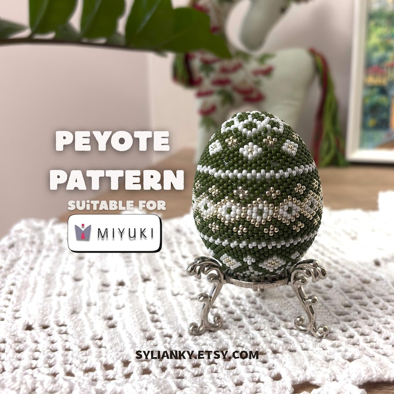 Może przedstawiać: Ozdobione koralikami jajko z zielonym, białym i złotym wzorem, prezentowane na dekoracyjnym srebrnym stojaku. Widoczny tekst "PEYOTE PATTERN" i "SUITABLE FOR MIYUKI". Jajko leży na białej koronkowej powierzchni.