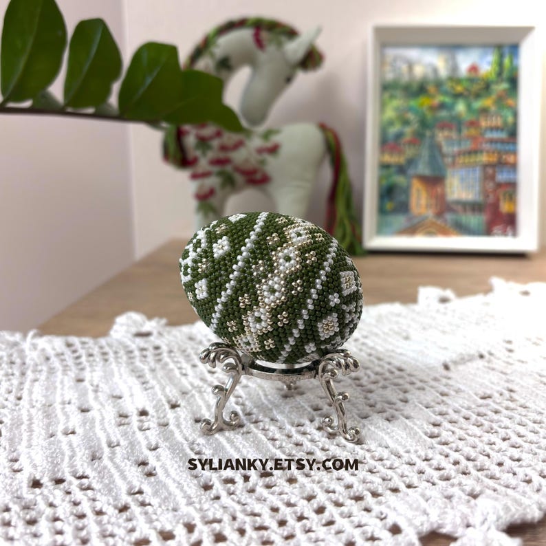 Może przedstawiać: Ozdobne jajko z koralik&oacute;w w odcieniach zieleni, bieli i złota, prezentowane na ozdobnym srebrnym stojaku. Jajko ma geometryczny wz&oacute;r. Biała koronka i oprawiony obraz są w tle. Widoczna jest nazwa sklepu Etsy, SYLIANKY.ETSY.COM.