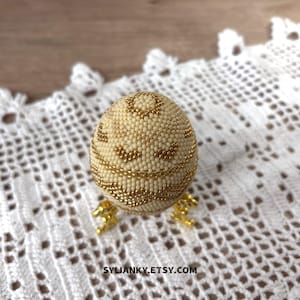 Puede incluir: Un huevo decorativo cubierto de peque&ntilde;as cuentas de color crema con detalles dorados. El huevo se apoya sobre un soporte dorado y est&aacute; colocado sobre una superficie blanca, similar al encaje. El nombre de la tienda Etsy, SYLIANKY.ETSY.COM, es visible.