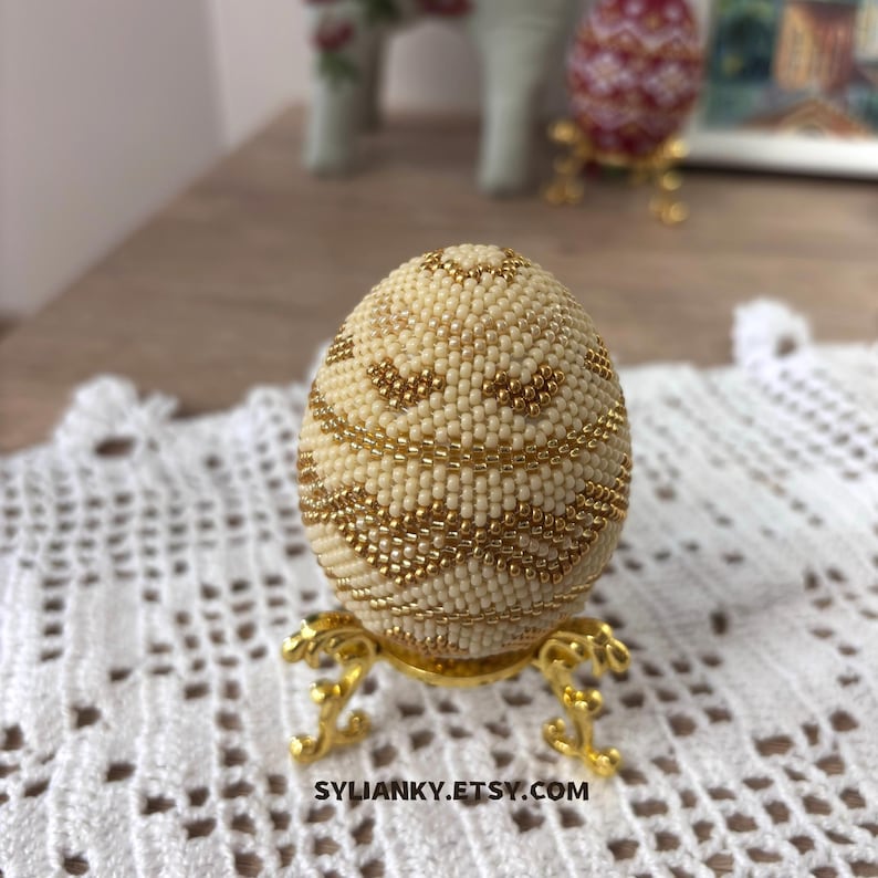 Puede incluir: Un huevo color crema con cuentas doradas, exhibido en un soporte dorado decorativo. El huevo presenta intrincados patrones de cuentas doradas. El nombre de la tienda Etsy, SYLIANKY.ETSY.COM, es visible en la parte inferior de la imagen.