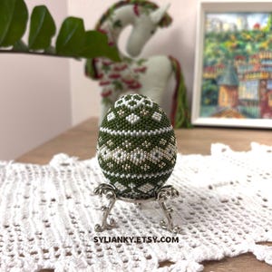 Może przedstawiać: Ozdobne jajko z koralik&oacute;w w odcieniach zieleni, bieli i srebra, prezentowane na srebrnym metalowym stojaku. Jajko ma geometryczny wz&oacute;r. Tło obejmuje białą koronkową serwetkę, pluszowego jednorożca i oprawiony obraz. Widoczna jest nazwa sklepu Etsy.