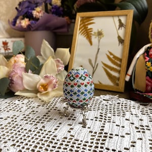 Peut inclure: Un &oelig;uf de P&acirc;ques perl&eacute; avec un motif g&eacute;om&eacute;trique dans des tons de bleu, vert, rouge et blanc. L'&oelig;uf est pr&eacute;sent&eacute; sur un support argent&eacute;.