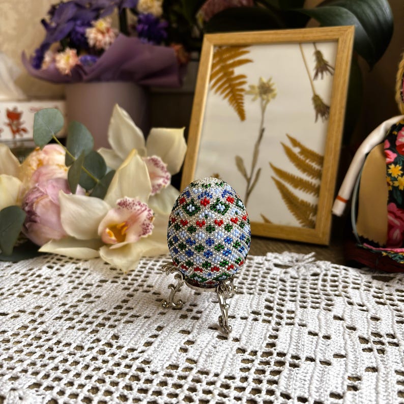 Peut inclure: Un &oelig;uf de P&acirc;ques perl&eacute; avec un motif g&eacute;om&eacute;trique dans des tons de bleu, vert, rouge et blanc. L'&oelig;uf est pr&eacute;sent&eacute; sur un support argent&eacute;.